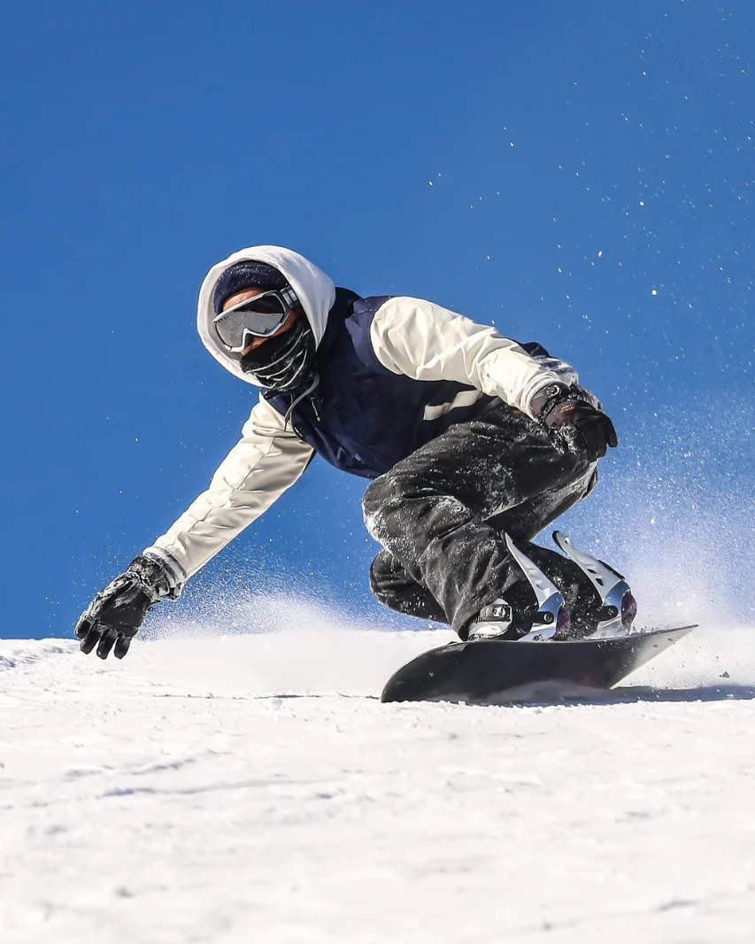 Snowboarder i hvid og sort jakke svinger aggressivt med sne der støver op under ren blå himmel