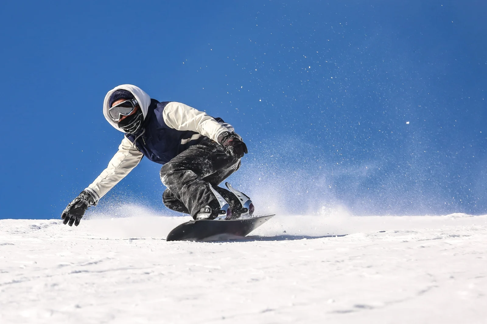 Snowboarder i hvid og sort jakke svinger aggressivt med sne der støver op under ren blå himmel